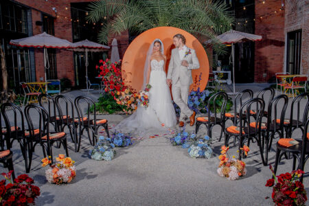 Bride and groom hold hands before an orange backdrop, with flowers and empty black chairs at an outdoor wedding ceremony.