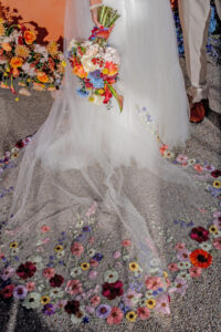 Bride in white dress with embroidered veil holds a vibrant bouquet; groom and bright flowers in background on gravel.