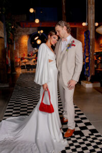 Bride in a white gown with red purse smiles by groom in beige suit on black-and-white checkered floor in a warmly lit venue.
