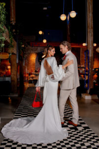 Bride in open-back white dress and groom in beige suit dance on checkered floor at a stylish, dimly lit venue.