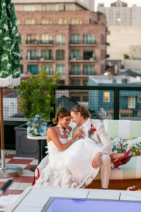 Bride in white dress and groom in light suit smile together on a rooftop lounge with city buildings in the background.