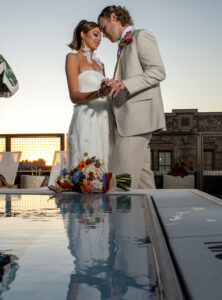 Bride in white dress and groom in beige suit hold hands by rooftop pool at sunset; bouquet rests on the ground.