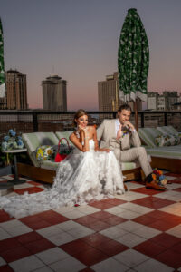 Bride in white gown and groom in beige suit sit on rooftop at sunset, city backdrop, green umbrellas, and red purse nearby.