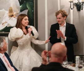 Woman in white dress speaks into mic as man applauds at joyful Louisiana wedding dinner; magnolia painting in background.