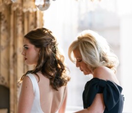 Bridesmaid in black assists bride in white gown beneath chandelier in elegant, sunlit Louisiana wedding room.
