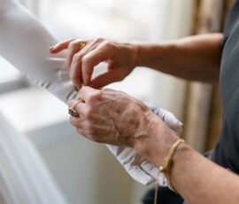 Older woman buttoning the sleeve of a white wedding dress by a window, capturing Gulf South wedding inspiration.