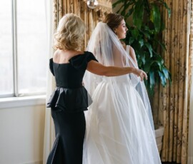 A bridesmaid in black helps a bride with her gown by a window, chandelier and plant highlighting a stunning Gulf South wedding.