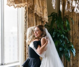 Bride in white dress hugs woman in black near gold curtains, both smiling during joyful New Orleans wedding ceremony.