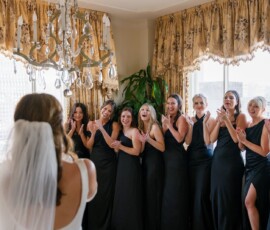 Bride in white dress and veil faces excited, smiling bridesmaids in black dresses by large windows at a Louisiana wedding.