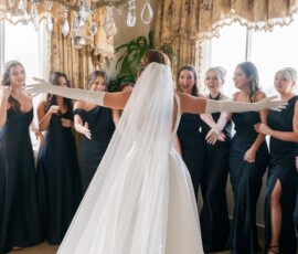 Bride in white gown and veil faces excited bridesmaids in black dresses, warmly lit room—ideal Louisiana wedding inspiration.