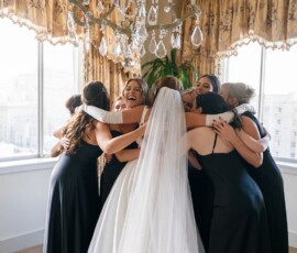 Bride in white gown hugs bridesmaids in black dresses beneath chandelier in bright New Orleans wedding venue.