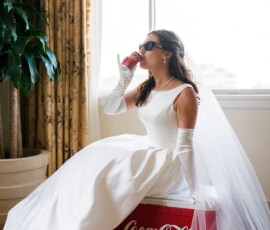 Bride in white dress and veil sips Coke on red cooler, wearing sunglasses and gloves; plant and window in background.