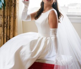 Bride in white dress and sunglasses smiles with Coca-Cola can, sitting on a red cooler by a window indoors—playful wedding vibe.