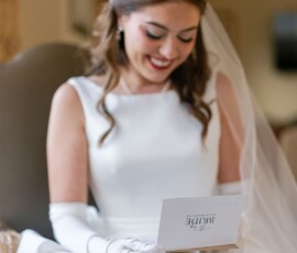 Bride with long brown hair and veil sits indoors in white dress, reading a BRIDE card before her Louisiana wedding ceremony.