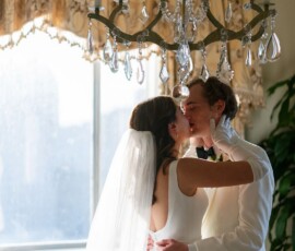 Bride and groom in white kiss under chandelier in sunlit room with window and elegant drapes, evoking Gulf South romance.