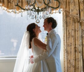 Bride and groom smiling and embracing under a chandelier in a sunlit, elegant New Orleans wedding venue with gold curtains.