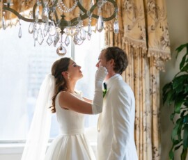 Bride in white dress and veil touches groom’s face as they smile in an elegant Louisiana wedding with golden curtains.