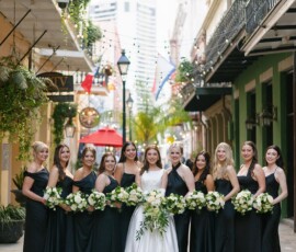 Bride in white gown with ten bridesmaids in black dresses, holding white and green bouquets on a New Orleans city street.