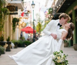Groom in tux dips and kisses bride in white gown with bouquet on a city street lit by string lights, New Orleans wedding.