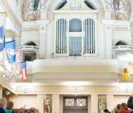 Bride in white walks down the aisle with two adults in a grand church, under chandeliers and a large pipe organ, as guests watch.
