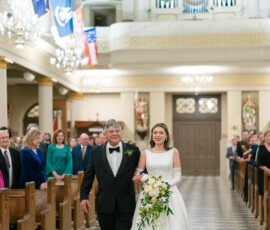Bride in white gown with bouquet walks down aisle with older man in tux as guests watch in decorated New Orleans church.