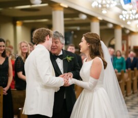 Bride in white dress smiles as her father greets the groom at a church wedding, with flags and chandeliers in the background.