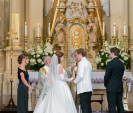 Bride and groom exchange rings at church altar with priest and witnesses, surrounded by gold accents, flowers, and candles.