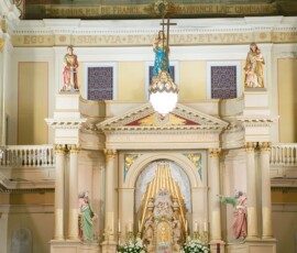 Bride and groom at ornate church altar with candles, statues, and flowers, as guests watch their wedding ceremony.