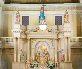 Bride and groom kneel at an ornate church altar with statues and mural as guests watch their Gulf South wedding ceremony.