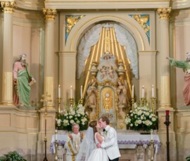 Bride and groom kiss at an ornate church altar during their wedding, with a priest and elegant statues in the background.