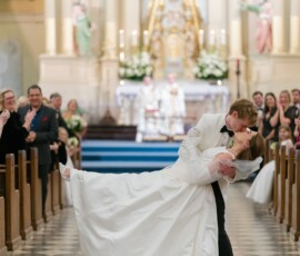 Groom in white tuxedo dips and kisses bride in church aisle at elegant New Orleans wedding, guests smiling in pews.