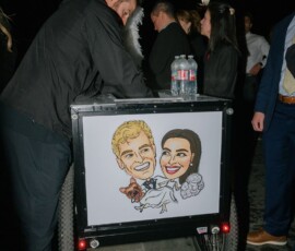 Man leans on a cart with a wedding caricature, dog, and feathers under a white umbrella; people enjoy the lively scene.