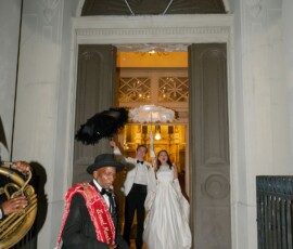 Newlyweds holding black and white umbrellas stand smiling in a doorway as a man leads with a banner; flags hang overhead.