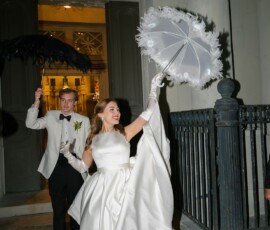 Bride in white gown and groom in white jacket walk at night with ornate umbrellas, joyful after New Orleans wedding.