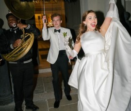 Bride in white gown dances with umbrella, joined by groom and sousaphone player at festive Louisiana wedding at night.