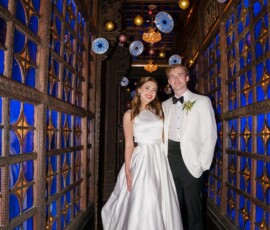 Bride and groom smiling in hallway with blue stained glass, ornate woodwork, and round lights at a New Orleans wedding.
