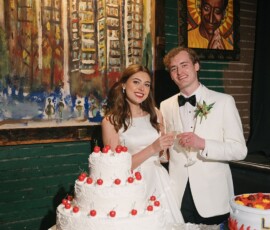 Bride and groom in white smile by a large wedding cake, holding glasses at their reception with colorful paintings behind.
