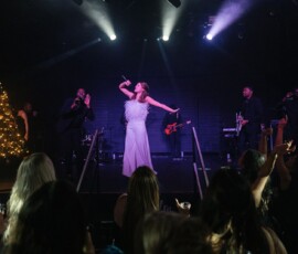 Female singer in white dress performs on stage with band, backup singers, audience, and a Christmas tree at a New Orleans wedding.