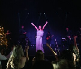 Bride in white dress raises arms on stage with band, guests cheer. Lit Christmas tree adds festive touch to lively wedding scene.