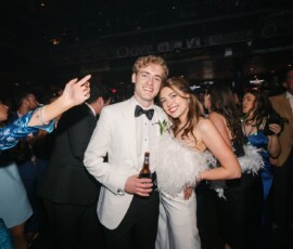 A smiling couple in white attire poses at a lively New Orleans wedding with dancing and celebration in the background.