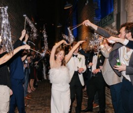 Bride in white dress dances under sparkling wands held by guests in a brick alleyway at night, celebrating her wedding.