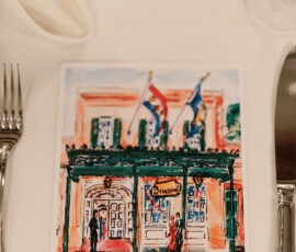 Colorful building illustration with three flags on a card, set between a fork and knife on a white tablecloth. Louisiana wedding.