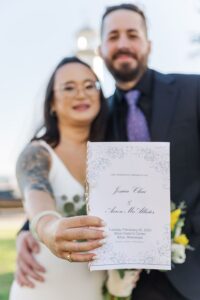 Smiling couple outdoors, woman holding wedding program for Jessica Chiu & Aaron McAllister, Biloxi, February 25, 2025.