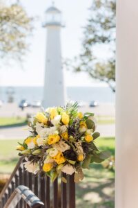 Yellow and white bouquet tied to a railing outdoors, with a white lighthouse against a blue sky in the background.