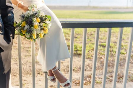 Bride in white dress on a railing, holding yellow and white bouquet, sunlight and greenery in background—Gulf South wedding.