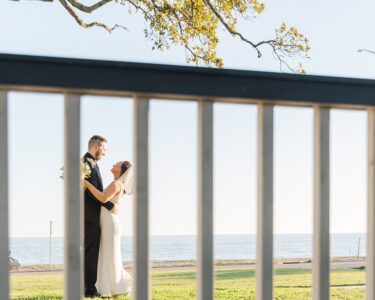 Bride and groom embrace by water, framed by a wooden fence and tree branch, in sunlit Louisiana wedding scene.