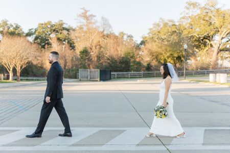 Bride in white dress and veil follows groom in black suit across crosswalk, bouquet in hand, classic Gulf South wedding scene.