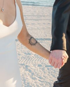 Couple holding hands on a sandy beach—a white dress, black suit, and tattoo—captures intimate seaside wedding inspiration.