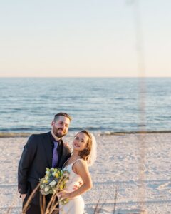 Bride and groom smile on a sandy beach after their ceremony, ocean and sky in the background, perfect for seaside wedding inspiration.