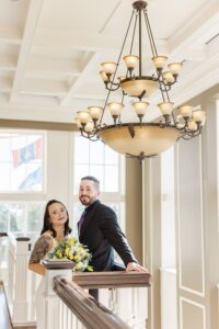 Formally dressed couple stands by an indoor railing at a New Orleans wedding, with flowers, large windows, chandelier, and flags visible.